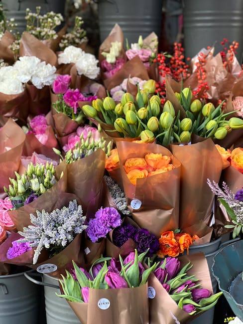 A vibrant display of freshly cut flowers arranged in brown paper wrapping, featuring a variety of blooms in multiple colours. Prominent are green tulip buds, white and purple lilacs, deep purple and p