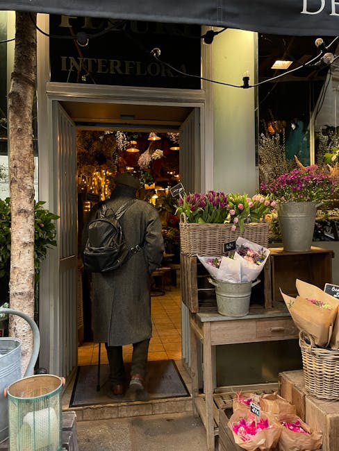 A person with a black backpack, dressed in a long gray coat, is seen entering a floral shop through a gray doorframe. Outside the shop, there is a display of fresh flowers in various containers, inclu