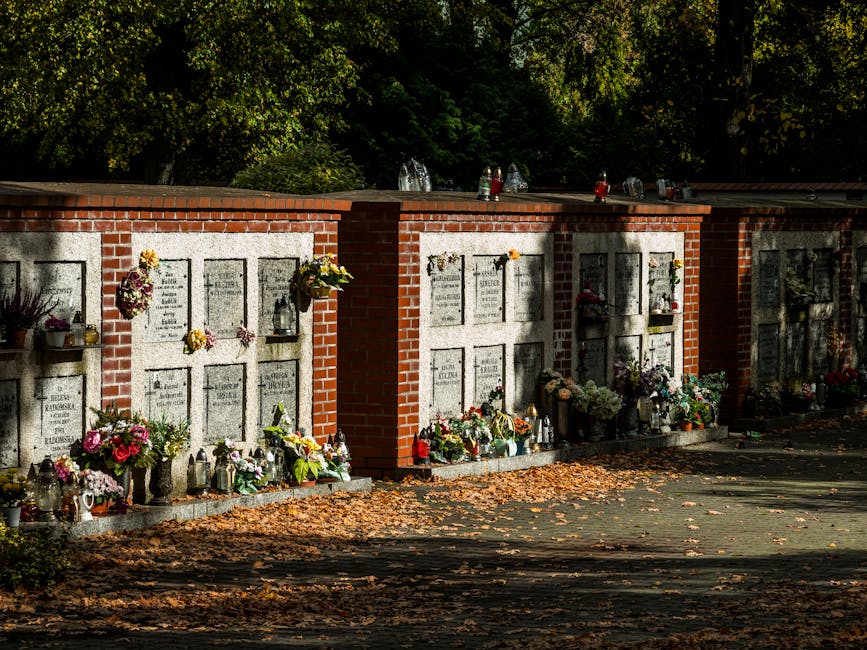 A row of cemetery memorial plaques mounted on a red brick wall, each adorned with various floral tributes including bouquets of pink, yellow, and white flowers, some in glass jars and vases, with addi