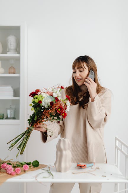 A woman with long brown hair and a beige sweater is holding a large, vibrant floral bouquet featuring red, white, and pink blooms, along with green foliage. She is smiling and speaking on a mobile pho