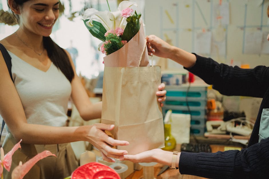 A female florist with long dark hair, wearing a light-colored sleeveless top, hands a wrapped bouquet of fresh white calla lilies, soft pink roses, and greenery to a customer. The bouquet is enclosed 