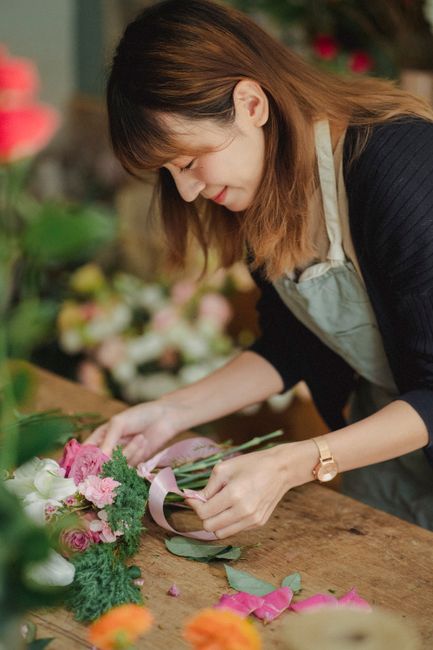 A female florist with brown hair, wearing a light gray apron over a dark long-sleeve shirt, is concentrating on arranging a bouquet of fresh flowers. She is holding a cluster of pink roses and greener