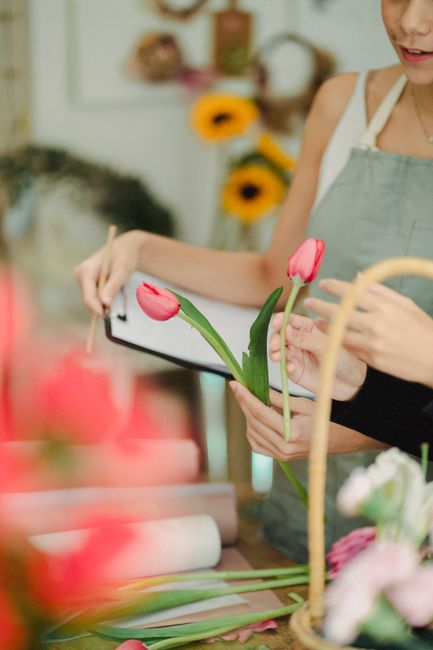 A person holding a smartphone in one hand and adjusting a vibrant pink tulip bouquet with their other hand inside a florist shop. The bouquet includes fresh, plump tulip petals and long green stems, a