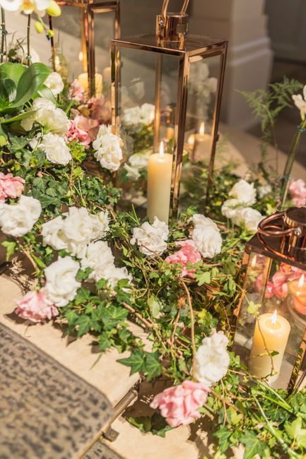 A floral arrangement featuring white and soft pink hydrangeas, interwoven with lush green ivy, forming a cascading display. The flowers are placed around elegant copper and glass lanterns with tall, l