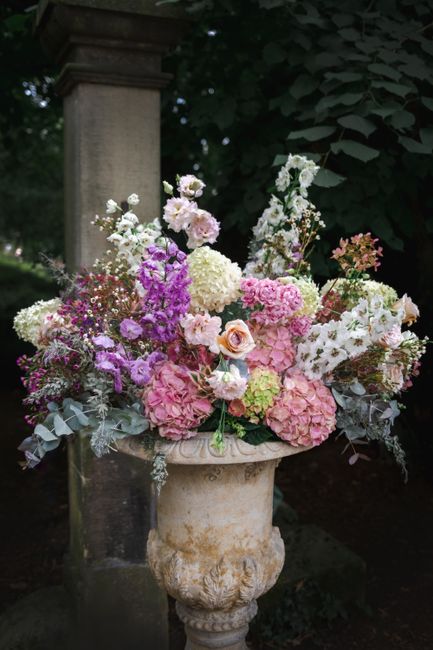 A large, lush floral arrangement placed in an elegant, weathered stone vase outdoors. The bouquet features a variety of fresh flowers including abundant pink hydrangeas, soft white and blush roses, de